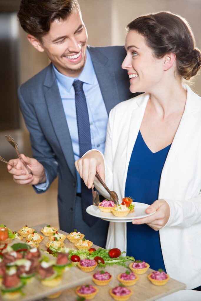 Happy young couple serving themselves with tartlets in buffet of restaurant, smiling at each other. Catering concept