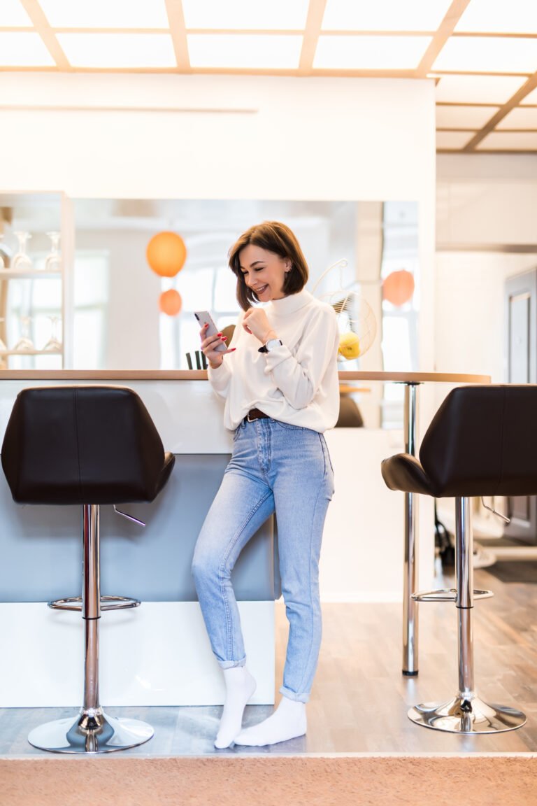 Girl with phone standing in panoramic kitchen with bright walls high table and bar chairs